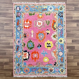 May include: A rectangular pink rug with a blue border and colorful floral patterns. The rug features a central pink field with various orange, yellow, and green floral designs. The border is light blue with intricate patterns.