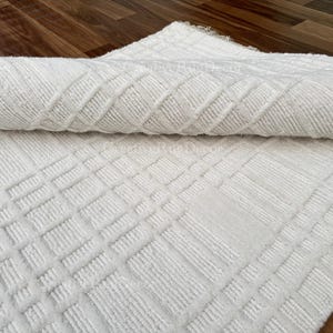 May include: A rolled-up white rug with a geometric pattern. The rug has a textured surface with a repeating square and diamond design. The rug is made of a soft, plush material, and is displayed on a wooden surface.