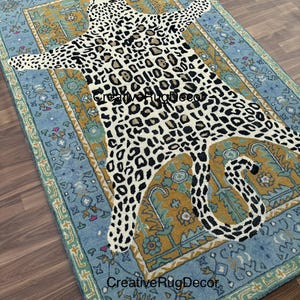 May include: A rectangular rug with a light blue border and a central design resembling a leopard skin. The leopard pattern is black and brown spots on a cream background. The rug has a decorative border with floral patterns.