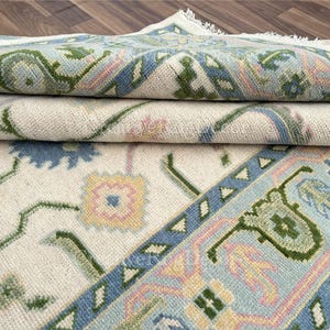 May include: Folded area rug with a cream-colored background and a detailed border. The border features a light blue, green, and pink design. The rug has a floral pattern with green stems and blue and yellow flowers.