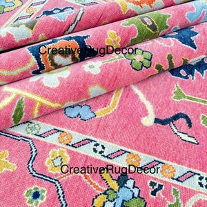 May include: Pink rug with a floral pattern in shades of blue, yellow, green, and orange. The rug has a border with a repeating pattern of lines and flowers.