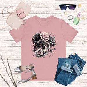 May include: A pink t-shirt with a black and white skull design surrounded by pink and black roses. The skull has intricate details and a spider web design.