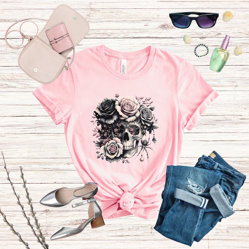 May include: Pink t-shirt with a black and white skull design surrounded by black and pink roses. The skull has a spider web design and a spider on the side.