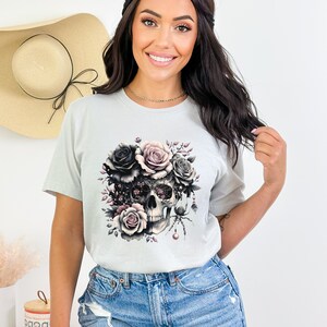 May include: A light gray t-shirt with a graphic print of a skull surrounded by black and pink roses. The skull has intricate details and a spider is perched on the top.