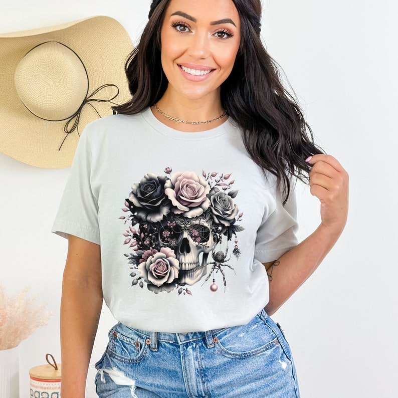 May include: A light gray t-shirt with a graphic print of a skull surrounded by black and pink roses. The skull has intricate details and a spider is perched on the top.