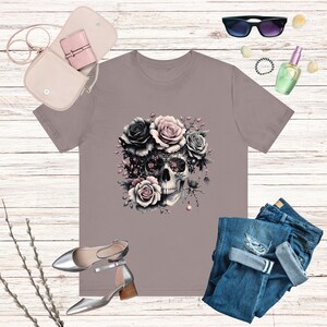 May include: A gray t-shirt with a skull design surrounded by pink and black roses. The skull has intricate details and is surrounded by a spider web.