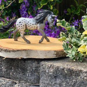 May include: A gray and white spotted horse figurine with a black mane and tail standing on a wooden slice. The figurine is surrounded by green foliage with purple flowers.