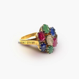 Op de afbeelding: Een gouden ring met een bloemvormig ontwerp met een cluster van gekleurde edelstenen, waaronder rood, blauw, groen en wit. De ring is gegraveerd met de woorden "MEERO JEWELS".