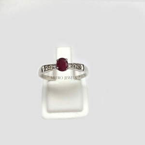 May include: A silver ring with a red ruby gemstone and small white diamonds on the band. The ring is on a white display stand. The text "MEERO JEWELS" is visible on the stand.