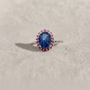Puede incluir: Anillo de plata con una gran gema de zafiro estrella azul oscuro de forma ovalada, rodeada de pequeñas gemas rosas redondas. El anillo tiene el texto "MEERO JEWELS" en el lateral.