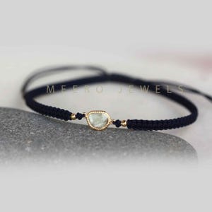 May include: A delicate bracelet with a light blue gemstone set in gold, tied with black cord. The bracelet is adjustable and features the text "MEERO JEWELS".