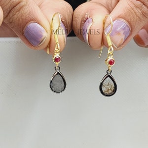 Op de afbeelding: Een paar gouden oorhangers met een traanvormige zwarte diamant en een klein rood edelsteentje bovenaan. De oorbellen worden vastgehouden door een hand.