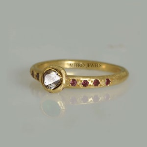 May include: A gold ring with a textured band and a single, round, grey diamond set in a bezel setting. The band is accented with small, red gemstones.