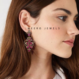 Op de afbeelding: Een paar oorhangers met een gouden zetting en rode edelstenen. De oorhangers zijn versierd met kleinere, heldere edelstenen. De oorhangers zijn gelabeld met "MEERO JEWELS".