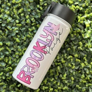 Pode incluir: Garrafa de água branca com um design rosa, roxo e branco que diz "Brooklyn Paige".