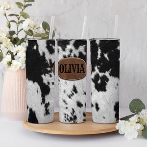 Könnte beinhalten: Drei Edelstahlbecher mit einem schwarz-weißen Kuhfellmuster. Der mittlere Becher hat einen braunen Lederfleck mit dem eingravierten Namen "Olivia".