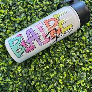 May include: A white stainless steel water bottle with a black lid and handle. The bottle has the word "BLAIRE" written on it in colorful, outlined letters.