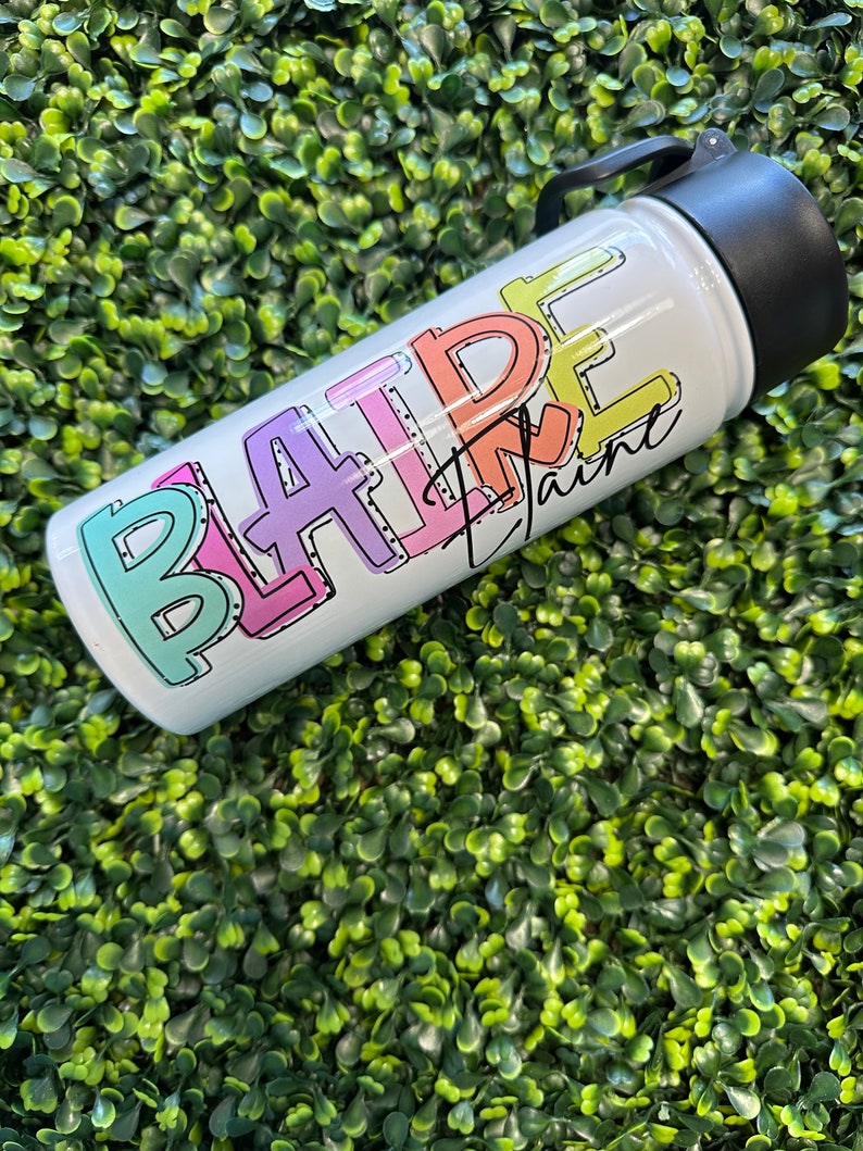 May include: A white stainless steel water bottle with a black lid and handle. The bottle has the word "BLAIRE" written on it in colorful, outlined letters.