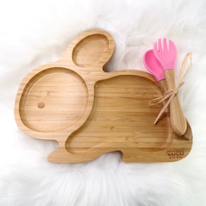 May include: A bamboo bunny-shaped plate with three compartments and a pair of pink and bamboo utensils. The plate has the text "coco and bro" on the bottom right corner. The utensils are tied together with a natural fibre.