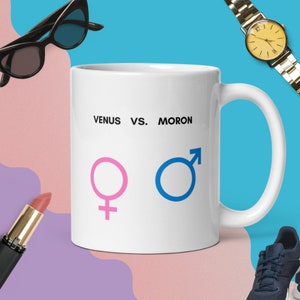 May include: White ceramic mug with the text "VENUS VS. MORON", a pink Venus symbol, and a blue Mars symbol. The mug is surrounded by a pair of black sunglasses, a gold watch, a tube of lipstick, and a pair of blue sneakers.