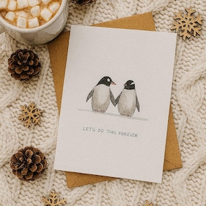 Könnte beinhalten: Grußkarte mit einer Pinguin-Illustration, die zwei Händchen haltende Pinguine zeigt, mit dem Text "LET'S DO THIS FOREVER." Die Karte liegt auf einem braunen Umschlag, umgeben von Tannenzapfen, Holzschneeflocken und einer Tasse heißer Schokolade.
