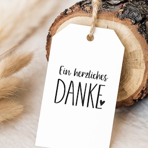 Könnte beinhalten: Ein weißes Geschenk-Etikett mit dem deutschen Satz "Ein herzliches DANKE" in schwarzer Schrift und einem kleinen Herz. Das Etikett hängt an einer natürlichen Schnur, vor einem Holzscheit und getrocknetem Laub.