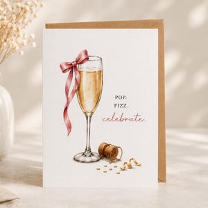 Könnte beinhalten: Eine weiße Grußkarte mit einer Aquarellillustration eines Champagnerglases gefüllt mit goldener Flüssigkeit, verziert mit einer altrosa Schleife. Unter dem Glas liegen ein Korken und goldenes Konfetti. Der Text lautet POP. FIZZ. celebrate.