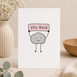 Puede incluir: Una tarjeta de felicitación blanca con una ilustración de dibujos animados de un personaje de roca gris con cara sonriente, brazos y piernas de palo negros, sosteniendo un letrero rectangular rosa con el texto escrito a mano you Rock en negro.