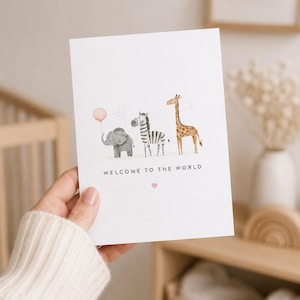 May include: A white greeting card featuring a watercolor illustration of a gray elephant holding a pink balloon, a black and white zebra, and a brown giraffe. The text "WELCOME TO THE WORLD" is printed in black below the animals with a small pink heart.