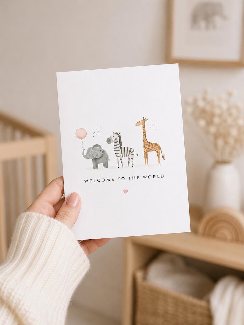 May include: A white greeting card featuring a watercolor illustration of a gray elephant holding a pink balloon, a black and white zebra, and a brown giraffe. The text "WELCOME TO THE WORLD" is printed in black below the animals with a small pink heart.