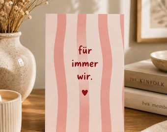 Puede incluir: Una tarjeta de felicitación rectangular con un patrón de rayas verticales onduladas de color rosa claro y blanco. El centro de la tarjeta muestra el texto rojo "für immer wir." seguido de un pequeño símbolo de corazón rojo debajo.