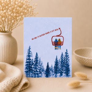 May include: A greeting card with a light blue background featuring a watercolour illustration of dark blue pine trees at the bottom. A red ski lift carries two figures, and the text reads: NO ONE I'D RATHER SHARE A LIFT WITH.