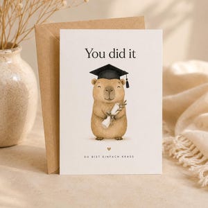 Peut inclure: Une carte de vœux blanche illustrée d'un capybara brun portant une toque de diplômé noire et tenant un diplôme enroulé. Le texte indique You did it et DU BIST EINFACH KRASS avec une petite icône de cœur entre les lignes de texte.