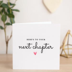 Könnte beinhalten: Eine weiße Grußkarte mit dem Text "HERE'S TO YOUR next chapter" in schwarzer Schrift, einem kleinen rosa Herz und einem minimalistischen Design. Die Karte liegt auf einer Holzoberfläche, mit einer Pflanze und dekorativen Gegenständen im Hintergrund.