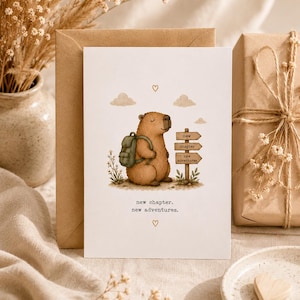 Può includere: Un biglietto d'auguri bianco con un capibara marrone che indossa uno zaino verde accanto a un cartello in legno con il testo new chapter new adventures. Il biglietto include piccole nuvole, fiori e il testo new chapter. new adventures.