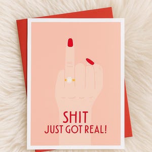 Può includere: Biglietto di auguri con una mano che fa il dito medio, smalto rosso e un anello d'oro con una perla. Il testo "SHIT JUST GOT REAL!" è stampato in rosso. Il biglietto è su una busta rossa.