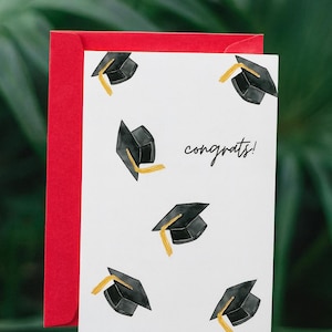 Könnte beinhalten: Eine weiße Abschlusskarte mit schwarzen Doktorhüten und goldenen Quasten, mit dem Wort "congrats!" in schwarzer Schrift. Die Karte wird mit einem roten Umschlag kombiniert, vor einem verschwommenen grünen Hintergrund.