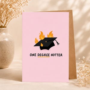 Könnte beinhalten: Eine hellrosa Grußkarte mit einer schwarzen Doktorhut-Illustration, die von orangefarbenen und gelben Flammen umgeben ist. Unter der Grafik steht der Text ONE DEGREE HOTTER in schwarzen Großbuchstaben mit einer gelben Linie unter degree.