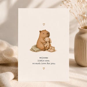 Könnte beinhalten: Eine Grußkarte mit einem braunen Wasserschwein, das ein kleines weißes Plüschkaninchen hält, daneben eine gefaltete Decke und Babyschuhe. Der Text lautet: welcome little one. so much love for you. Kleine braune Herzen befinden sich über und unter dem Motiv.