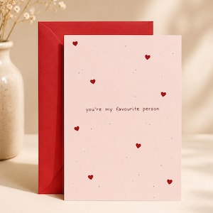 Pode incluir: Um cartão de felicitações rosa claro com pequenos corações vermelhos e pontos pretos espalhados pela superfície. O texto "you're my favourite person" está impresso no centro. Um envelope vermelho liso está posicionado atrás do cartão.