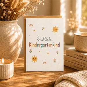 May include: A white greeting card featuring the German text Endlich Kindergartenkind in a mix of dark grey and colourful letters, surrounded by illustrations of smiling suns, small rainbows, flowers, and stars on a plain white background.