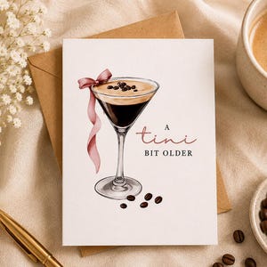Peut inclure: Une carte de vœux blanche avec une illustration à l'aquarelle d'un verre à martini rempli de café noir, surmonté de trois grains de café et d'un ruban rose. Le texte indique A tini BIT OLDER en lettres noires et roses.