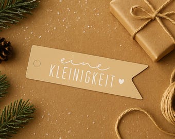 Könnte beinhalten: Ein beiger Geschenkanhänger mit dem Schriftzug "eine Kleinigkeit" in Weiß, einem kleinen Herz und einem Loch. Auf braunem Untergrund mit Geschenk, Bindfaden, Tannennadeln und einem Tannenzapfen.