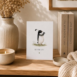 Peut inclure: Une carte de vœux blanche avec une illustration à l'aquarelle d'un manchot tenant un club de golf sur un brin d'herbe verte. Le texte GO FOR IT! est imprimé en noir sous le manchot, suivi d'une petite icône de cœur de couleur verte.