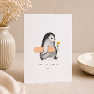 May include: A white greeting card featuring a charcoal-style illustration of a small penguin carrying a beige adhesive bandage and a tiny glass vase with a single yellow tulip. The text "FEEL BETTER SOON" is printed below with a small heart.