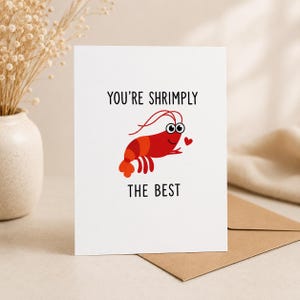 Könnte beinhalten: Eine weiße Grußkarte mit einer roten Cartoon-Garnele mit großen Augen und einem kleinen roten Herz. Der Text lautet YOU'RE SHRIMPLY THE BEST in schwarzen Großbuchstaben. Die Karte liegt neben einem braunen Umschlag auf einer hellen Fläche.