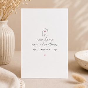 Peut inclure: Une carte blanche avec l'inscription manuscrite « new home, new adventures, new memories ». Un dessin de maison avec un cœur est au-dessus du texte. La carte est posée sur une surface neutre, avec un vase de fleurs séchées.