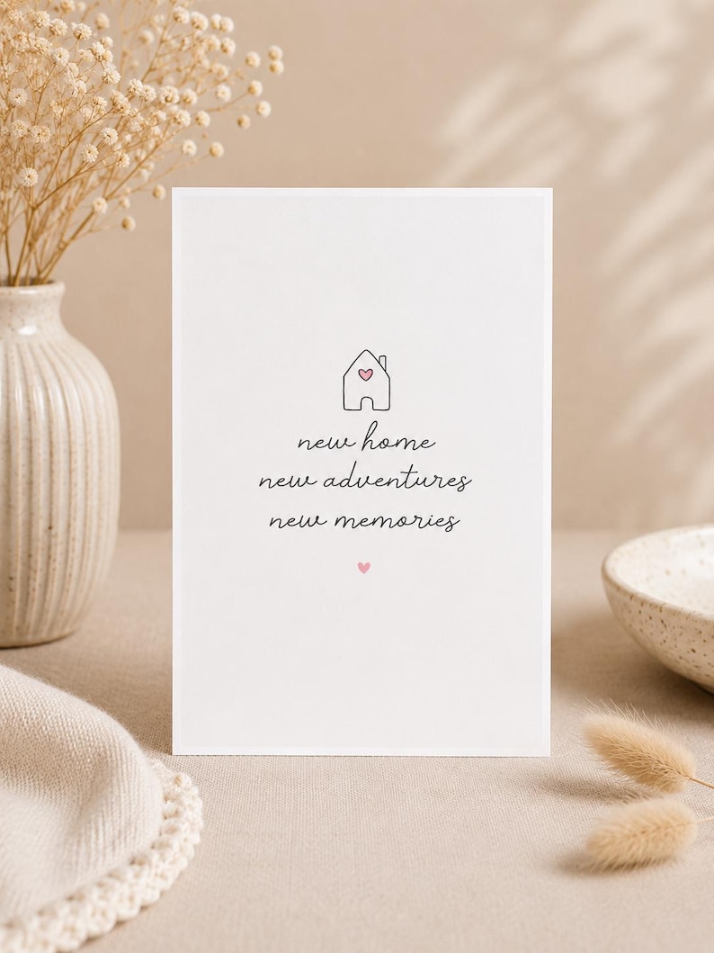 May include: A white card with the words "new home, new adventures, new memories" in a handwritten font. A small house graphic with a heart is above the text. The card is on a neutral surface with a vase of dried flowers.