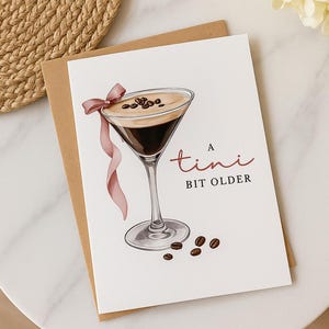 Puede incluir: Tarjeta de felicitación con una ilustración de martini. La copa contiene un líquido oscuro, cubierto con espuma y granos de café, y una cinta rosa. El texto dice "A tini bit older".