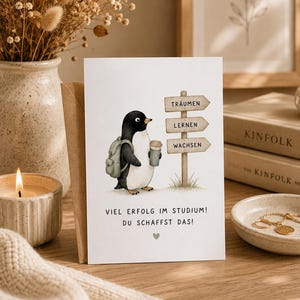 Könnte beinhalten: Eine Grußkarte mit der Illustration eines Pinguins, der einen Rucksack trägt und einen Kaffeebecher hält, neben einem Wegweiser mit den Wörtern TRÄUMEN, LERNEN, WACHSEN. Darunter steht VIEL ERFOLG IM STUDIUM! DU SCHAFFST DAS! mit einem Herz.